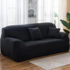 A modern black stretch sofa cover placed in a bright living room with sheer white curtains and a wooden floor.