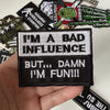 A hand holds a black iron-on slogan patch that reads I'M A BAD INFLUENCE BUT DAMN I'M FUNIII.