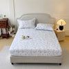 A white floral printed cotton fitted sheet is displayed on a bed with a light gray upholstered headboard.