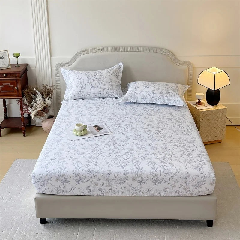 A white floral printed cotton fitted sheet is displayed on a bed with a light gray upholstered headboard.