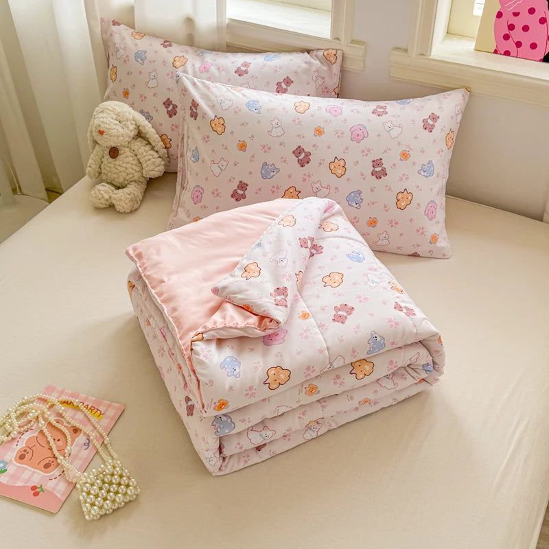 A pink and white quilted blanket with a floral pattern is displayed on a light-colored surface next to a stuffed animal.