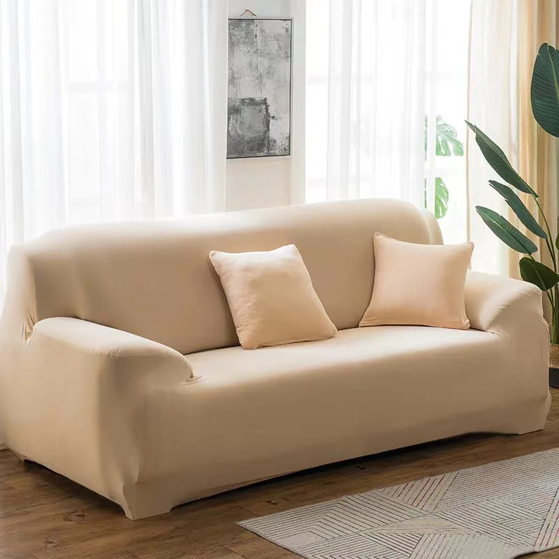 A beige stretch sofa cover with two light pink pillows placed on a modern sofa in a bright living room.