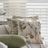 A close-up of a cream floral cushion cover with embroidered daisies and green foliage resting on a light-colored sofa.