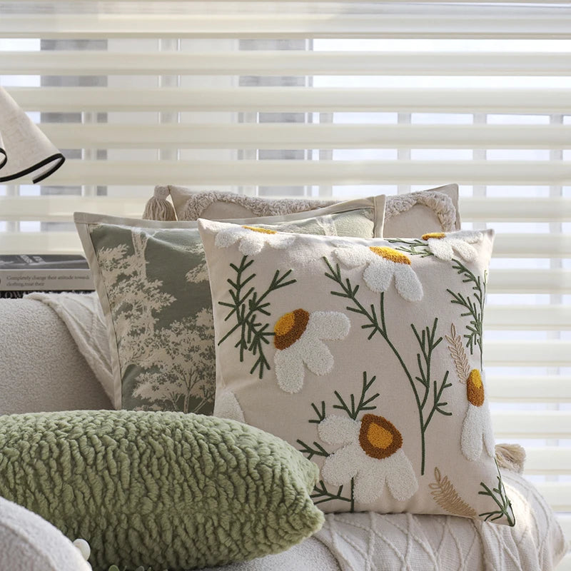 A close-up of a cream floral cushion cover with embroidered daisies and green foliage resting on a light-colored sofa.