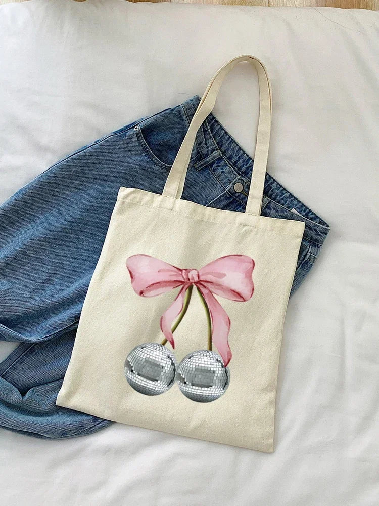 A beige canvas shopping bag featuring a pink bow tied around a pair of silver, spherical earrings.