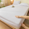 A hand is touching a white fitted bed sheet on a bed, showing the texture and fit of the fabric.