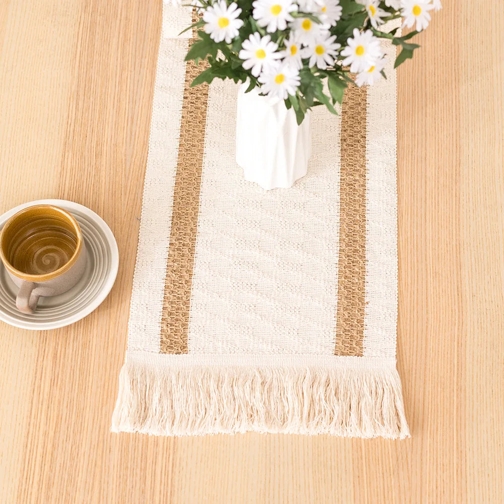 A beige linen cotton table runner with gold stripes and tassels, featuring a white flower arrangement.