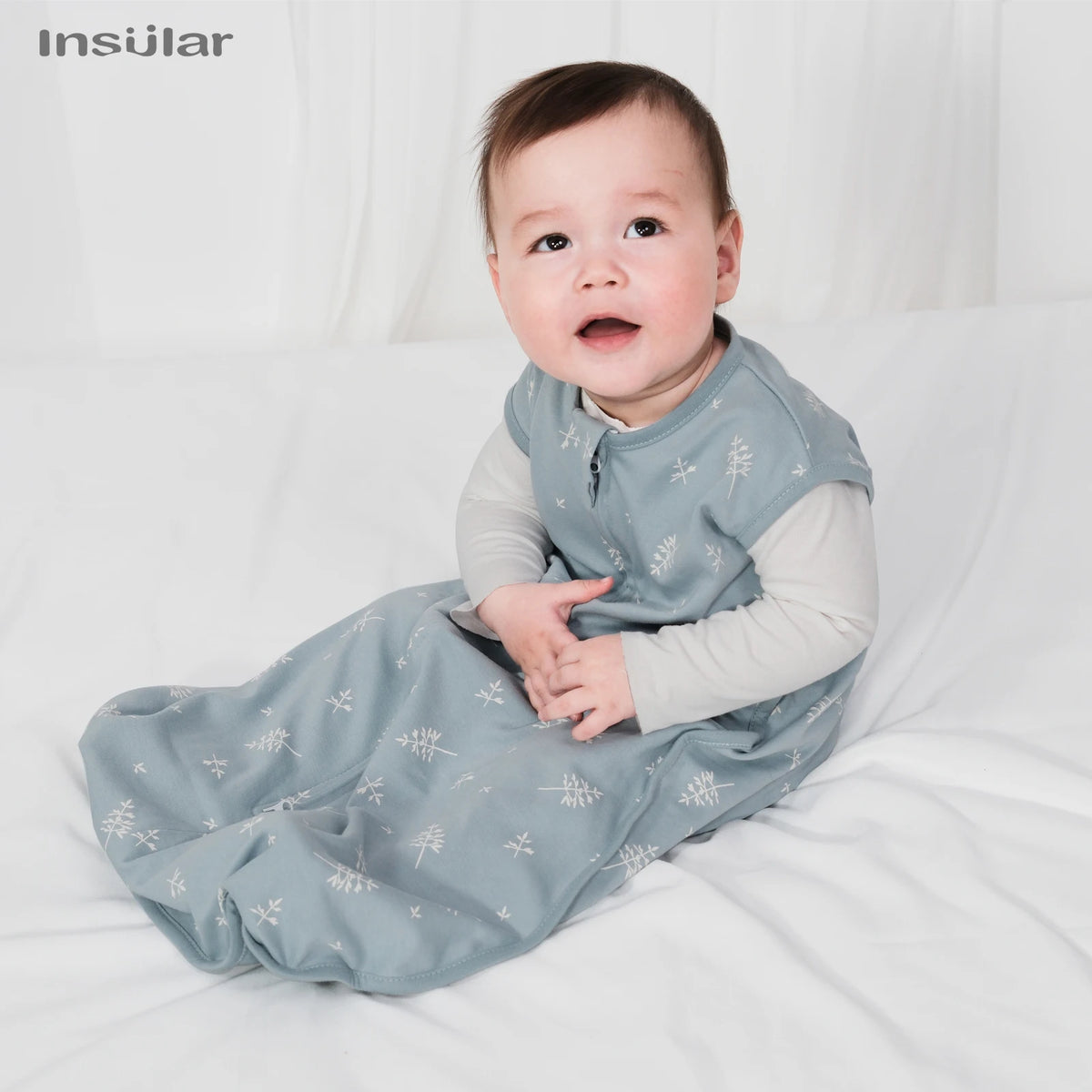 A baby wearing a light blue patterned cotton sleep sack is lying on white bedding.