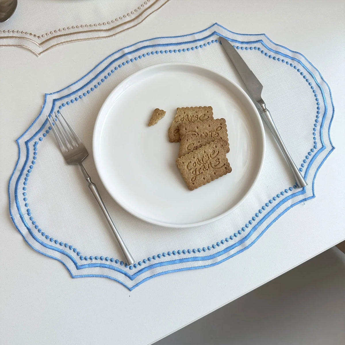 A white plate with three crackers is set on a blue embroidered linen placemat with a silver knife.