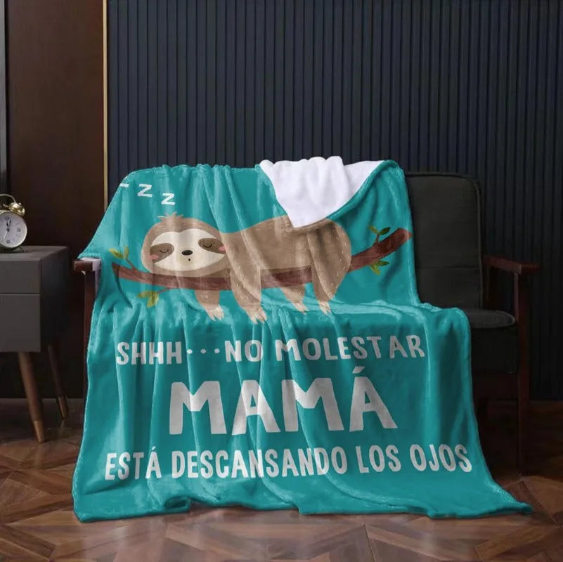 A teal knit throw blanket featuring a sloth design and Spanish text that reads 'SHHH NO MOLESTAR MAMÁ ESTÁ DESCANSANDO LOS OJ