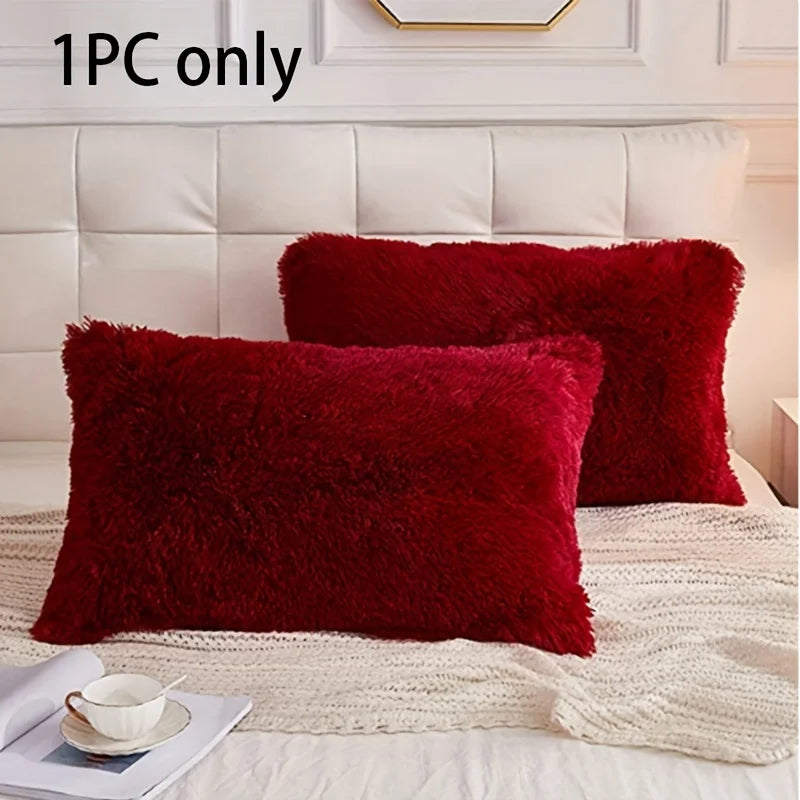 A single deep red faux fur pillow resting on a cream textured blanket in a bright bedroom setting.