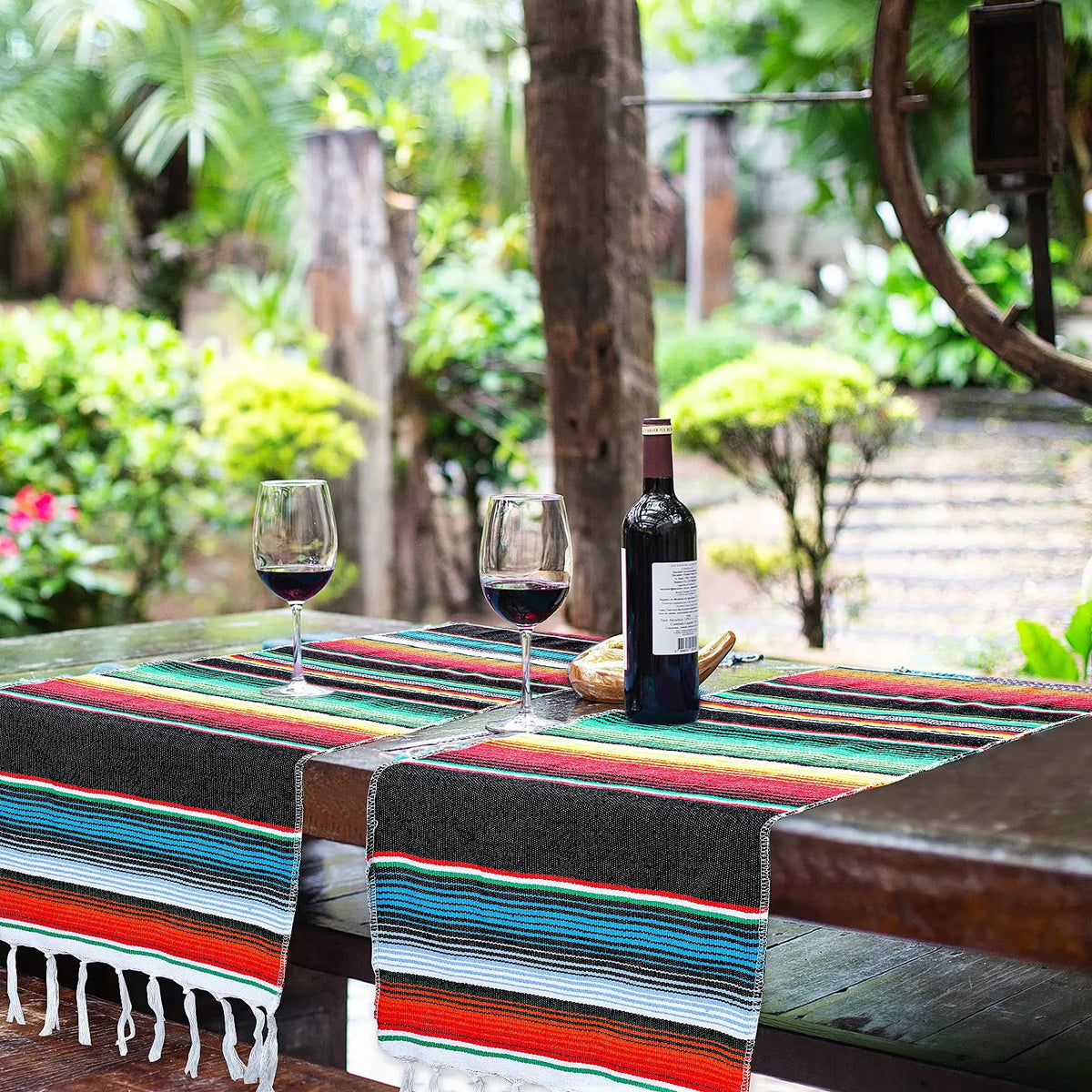 A dark bottle of wine sits on a striped Mexican serape table runner on a wooden table outdoors.