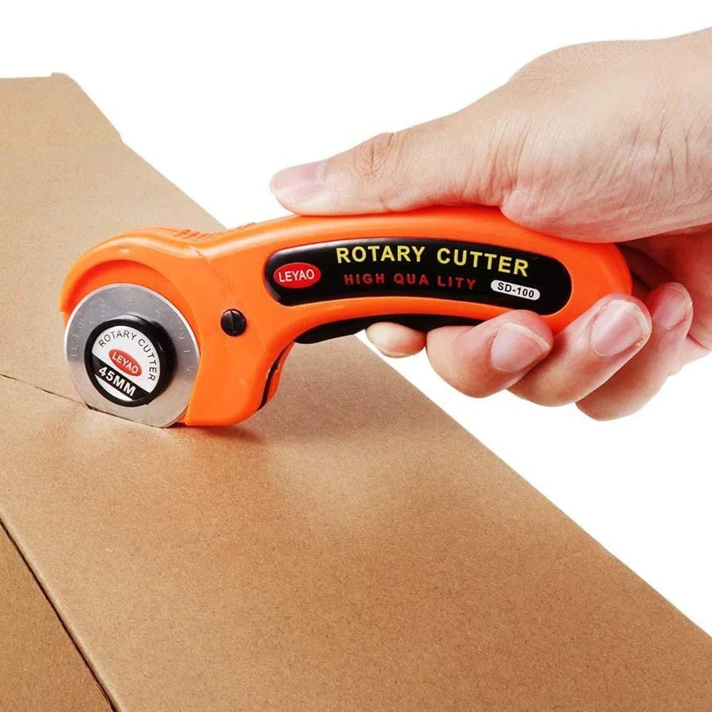 A hand holds an orange rotary cutter tool with a black handle and silver blade on brown cardboard.