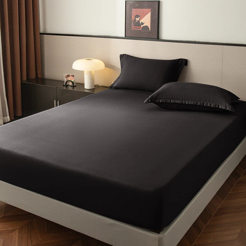 A dark gray fitted sheet is laid on a bed, next to a bedside table with a lamp and framed art.
