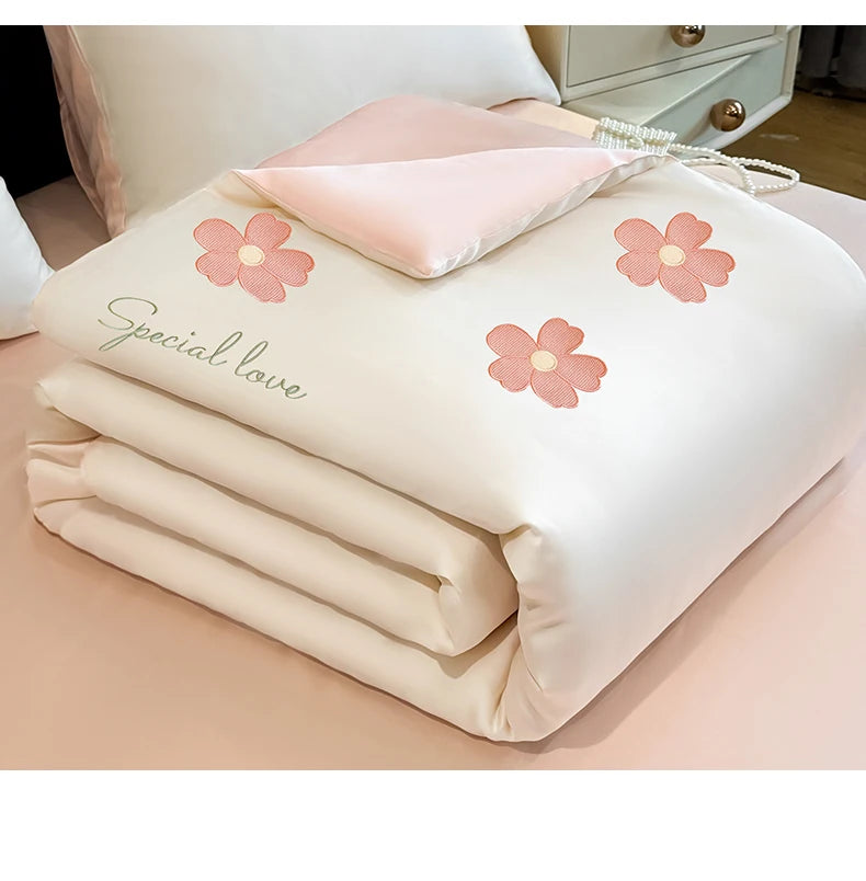 A white embroidered quilt with pink floral designs and a pink pillow, rolled up on a light pink surface.