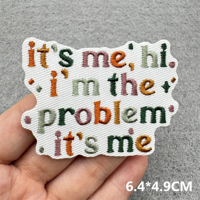 A hand holds a white embroidered patch with the text it's me hi i'm the problem it's me