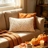 A cozy living room scene featuring a beige sofa with an orange throw blanket and a white embroidered leaf pillow.