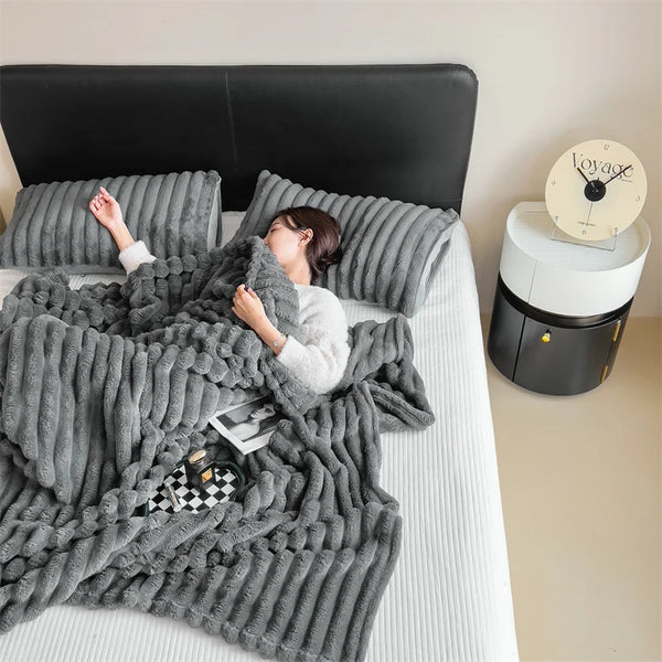A woman is relaxing on a bed covered with a gray plush winter blanket and a black headboard.