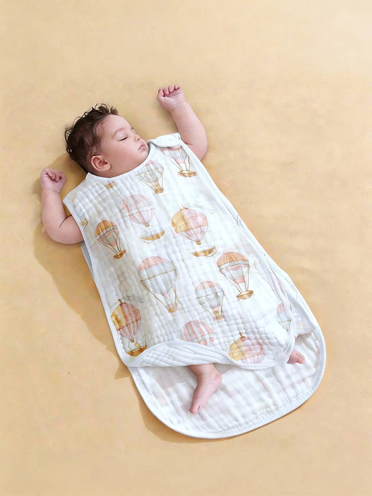 A baby is sleeping on a white fabric sleeping bag patterned with colorful hot air balloons.
