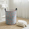 A light blue and pink patterned foldable canvas laundry basket sitting on a woven mat next to sheer white curtains.