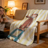 A yellow and white striped plush throw blanket featuring a cartoon illustration of C-Curious George sitting on a blue stool.