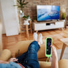 A hand holds a lime green silicone remote cover with a lanyard, resting on a couch.