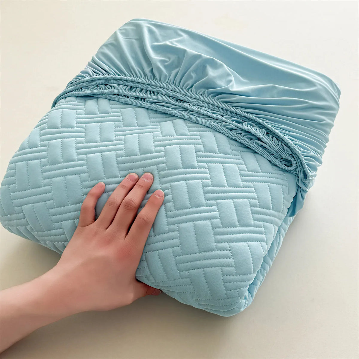 A hand is holding a light blue cooling spandex fitted sheet mattress protector with quilted texture.