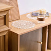 A set of four round, cream-colored embroidered placemats are displayed on a light wood table next to a wooden cabinet.