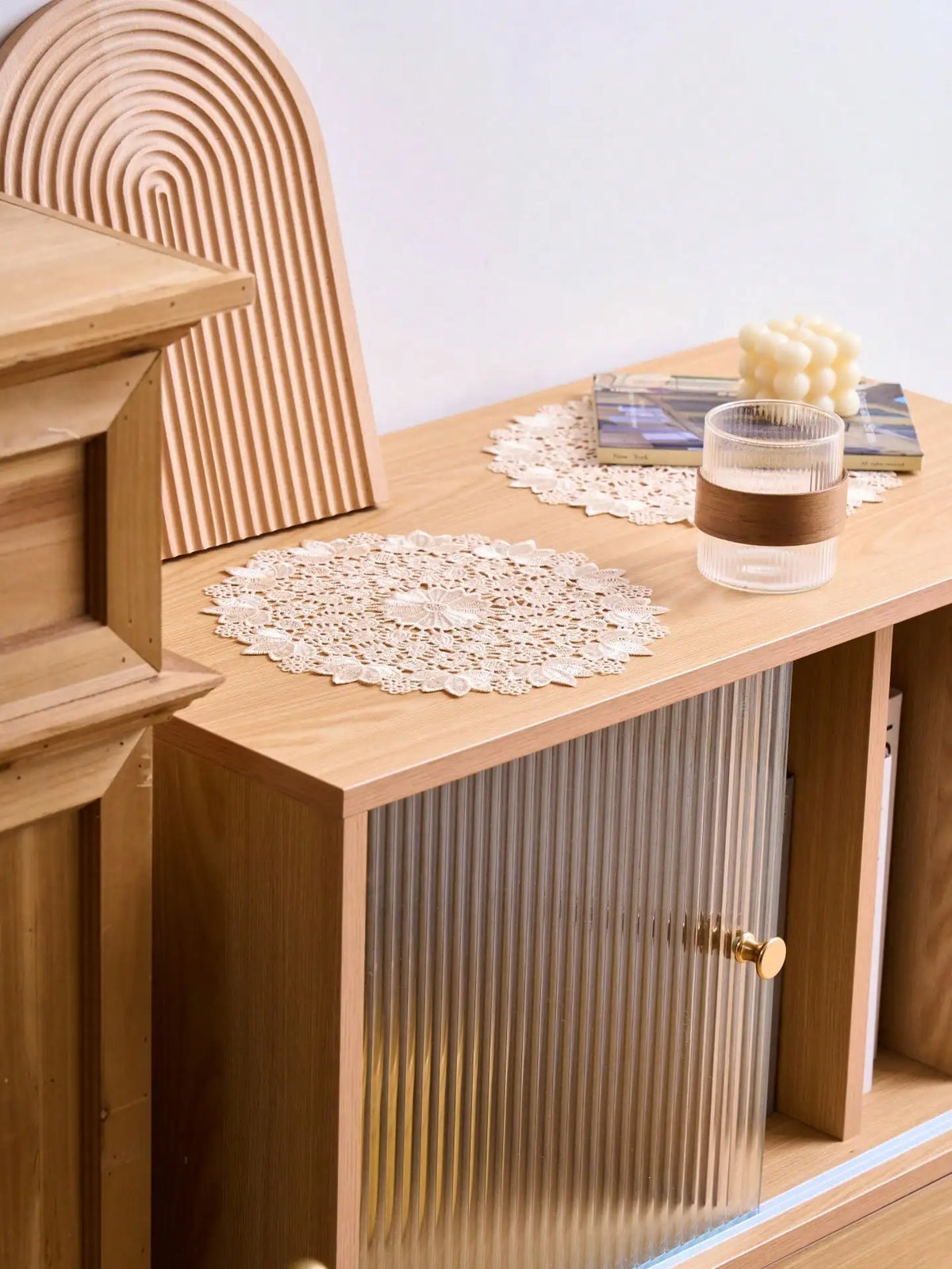 A set of four round, cream-colored embroidered placemats are displayed on a light wood table next to a wooden cabinet.