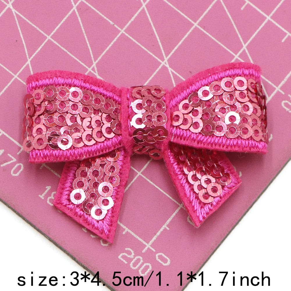 Pink sequin bow patches for DIY decoration displayed on a pink grid background.