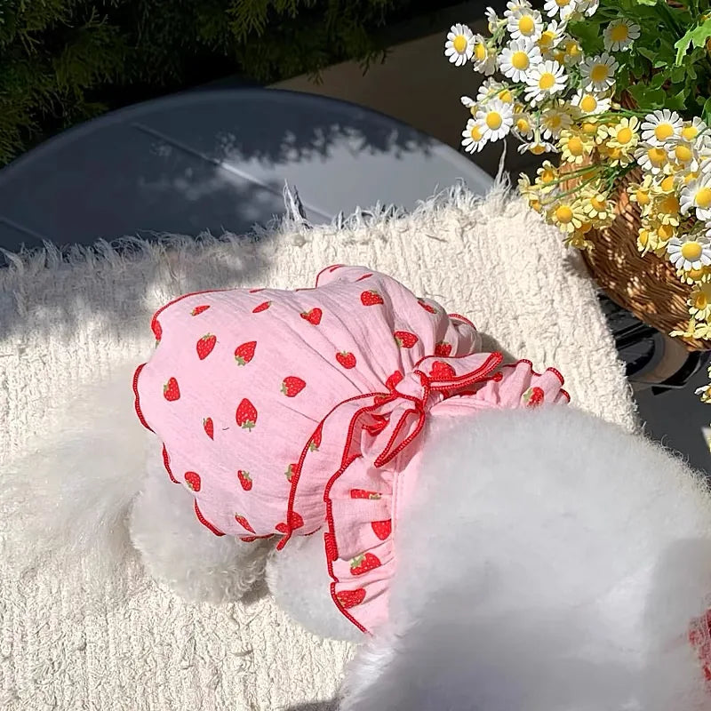 Pink strawberry print dog dress displayed on a white fluffy surface with yellow flowers in the background