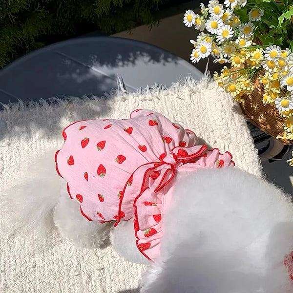 Pink strawberry print dog dress displayed on a white fluffy surface with yellow flowers in the background