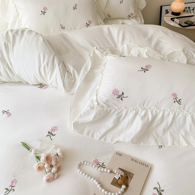 A white embroidered rose duvet cover set displayed on a bed with soft lighting and a magazine.