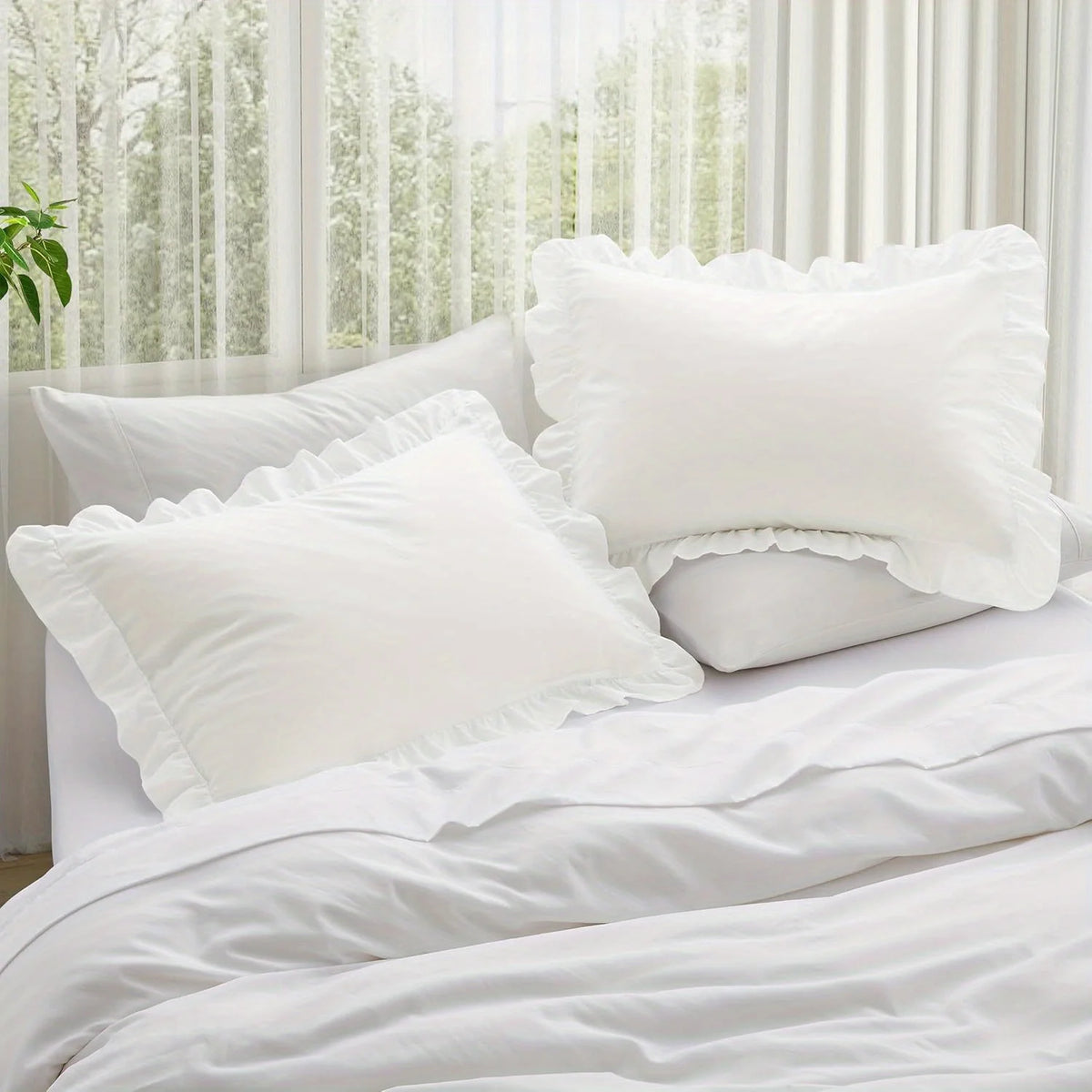 A set of two white ruffled envelope closure pillow covers resting on a bed with sheer curtains in the background.