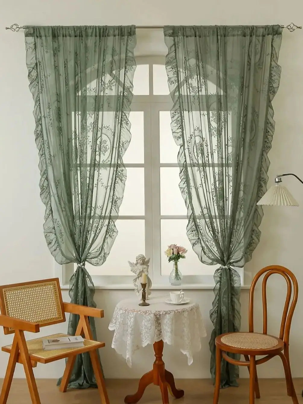 Pair of sheer sage green lace curtains framing a window with a white table and chairs.