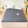 A gray quilted mattress protector cover placed on a white bed with white pillows and a small cup.