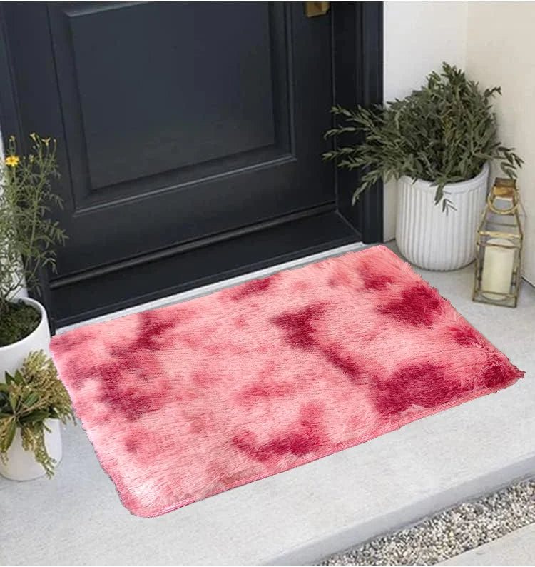 A rectangular pink and red patterned area rug placed on a light gray floor next to a dark door and potted greenery.