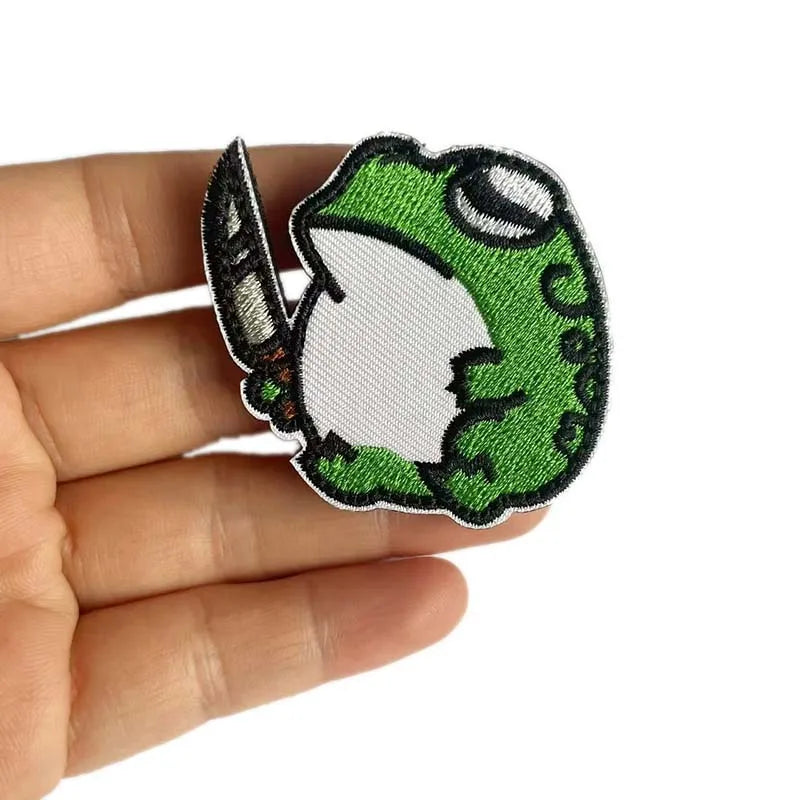 A hand holds a green and white animal embroidery patch featuring a stylized frog or lizard design.