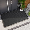 A black non-slip rubber mat is placed on a tiled floor outside a gray door with a potted plant nearby.