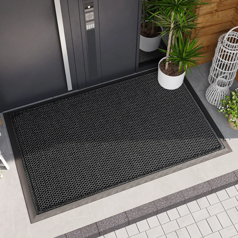 A black non-slip rubber mat is placed on a tiled floor outside a gray door with a potted plant nearby.