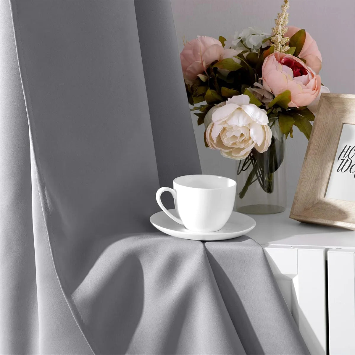 A white cup rests on a table next to a vase of pink and white flowers, with a gray curtain draped in the background.