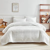White quilted cotton bedspread set with matching pillows on a bed against a white wall.