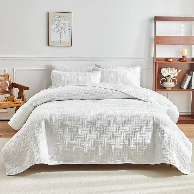 White quilted cotton bedspread set with matching pillows on a bed against a white wall.