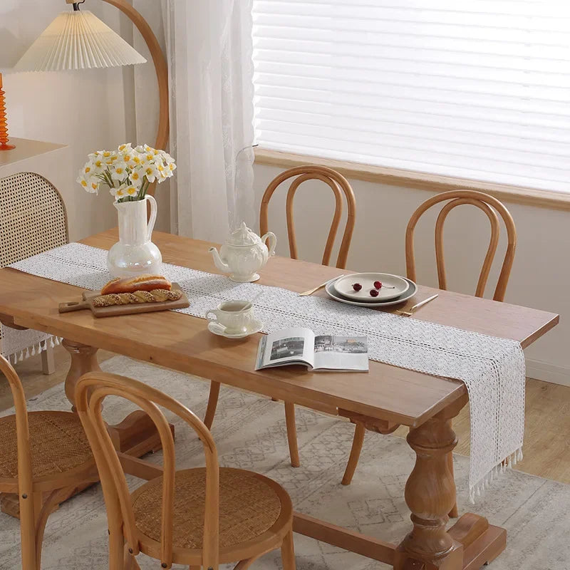 A wooden dining table set with a white crochet lace table runner and fringe, surrounded by wooden chairs.