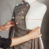 Hands are sewing a plaid dress using nickel-plated pins with colorful heads on a white mannequin.