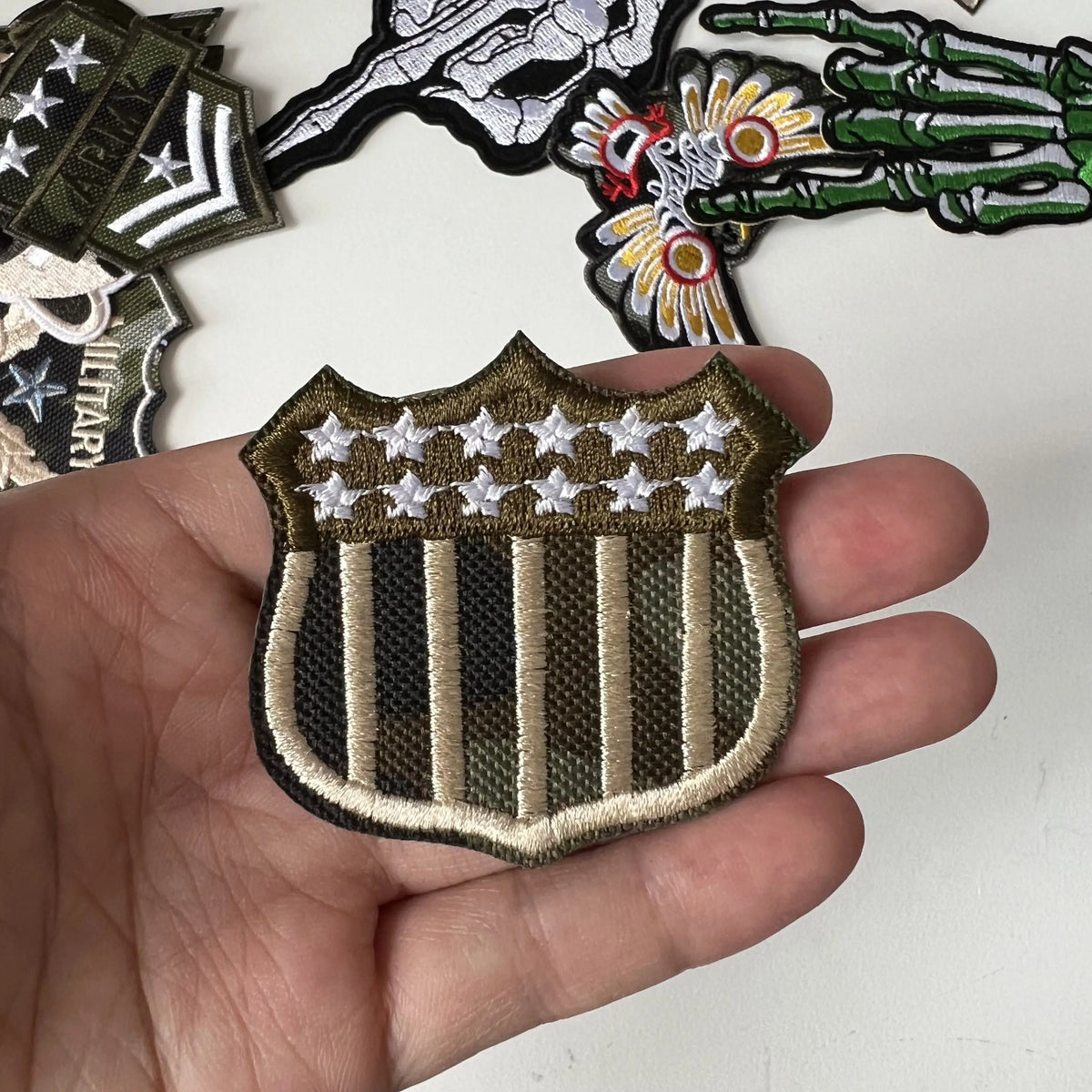 A hand holds a military-themed embroidered patch featuring vertical stripes and stars on a dark green background.