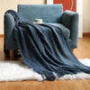 A dark blue knitted tassel throw blanket draped over a teal armchair next to a cup and saucer.