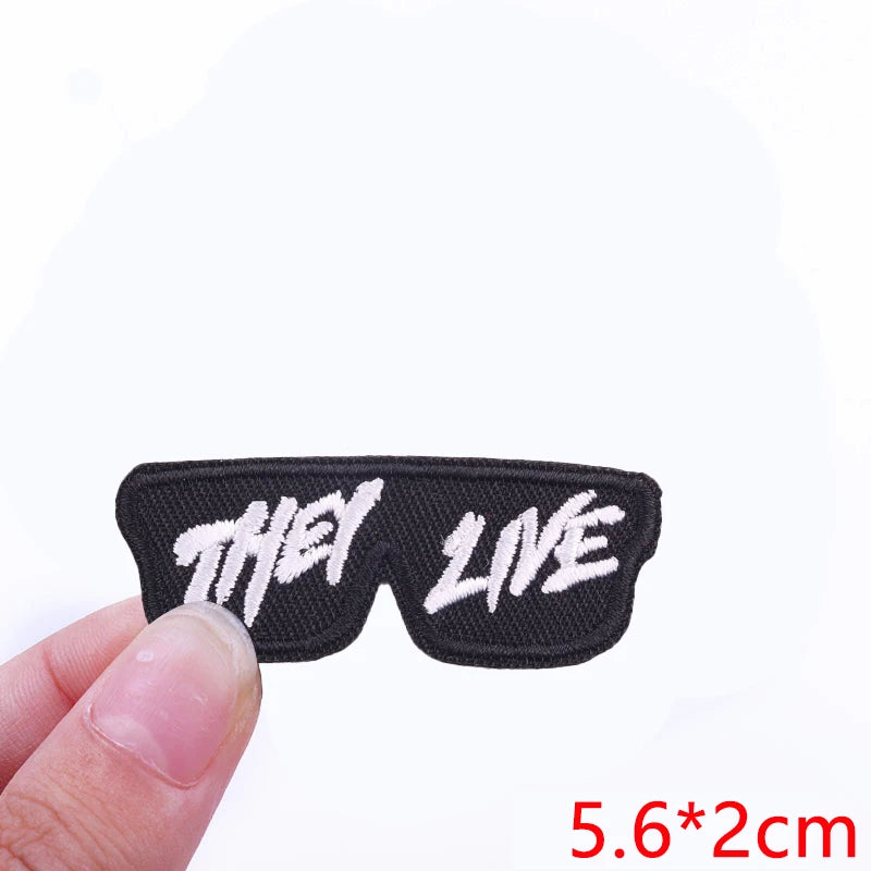 Black embroidered patch with the words THEY LIVE on a finger