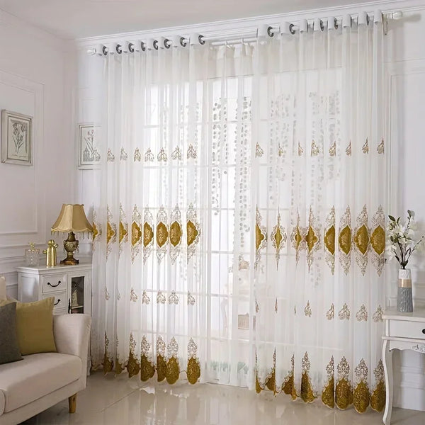 White sheer gauze curtain with gold embroidery covering a glass door in a bright room.