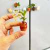 A hand holds a small terracotta ceramic vase with green leaves and small flowers against a white refrigerator door.